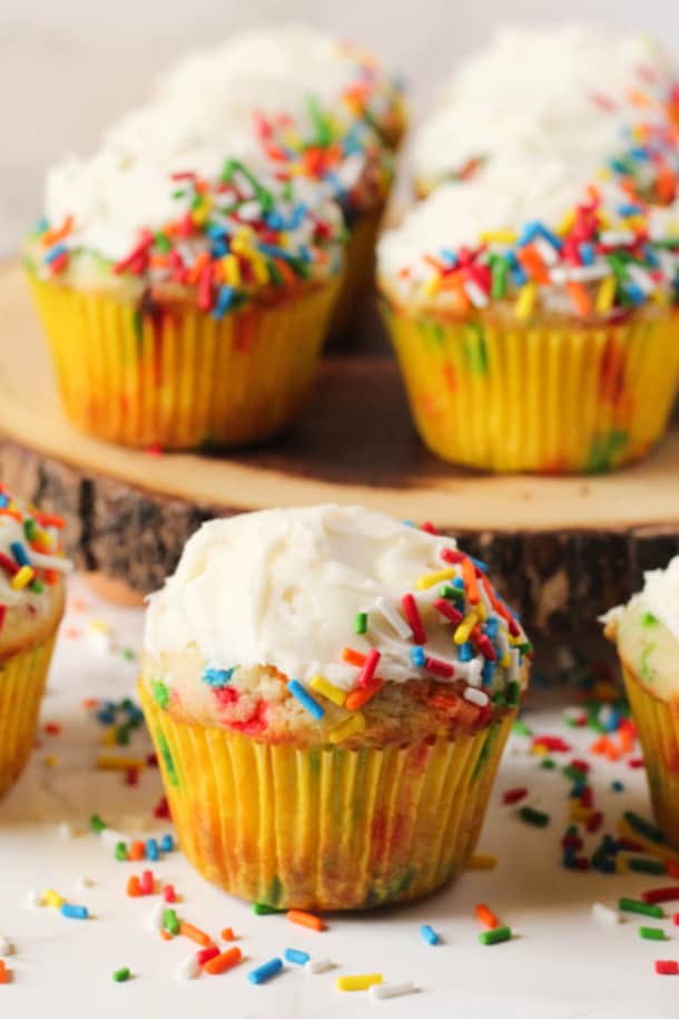 Gluten-free Funfetti Cupcakes on a wood tray, with one cupcake up close, with sprinkles all around