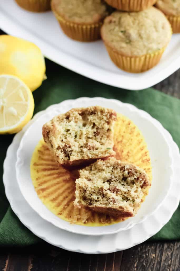 A Lemon Zucchini Oat Muffin cut in half so you can see inside, on a yellow cupcake liner and two plates