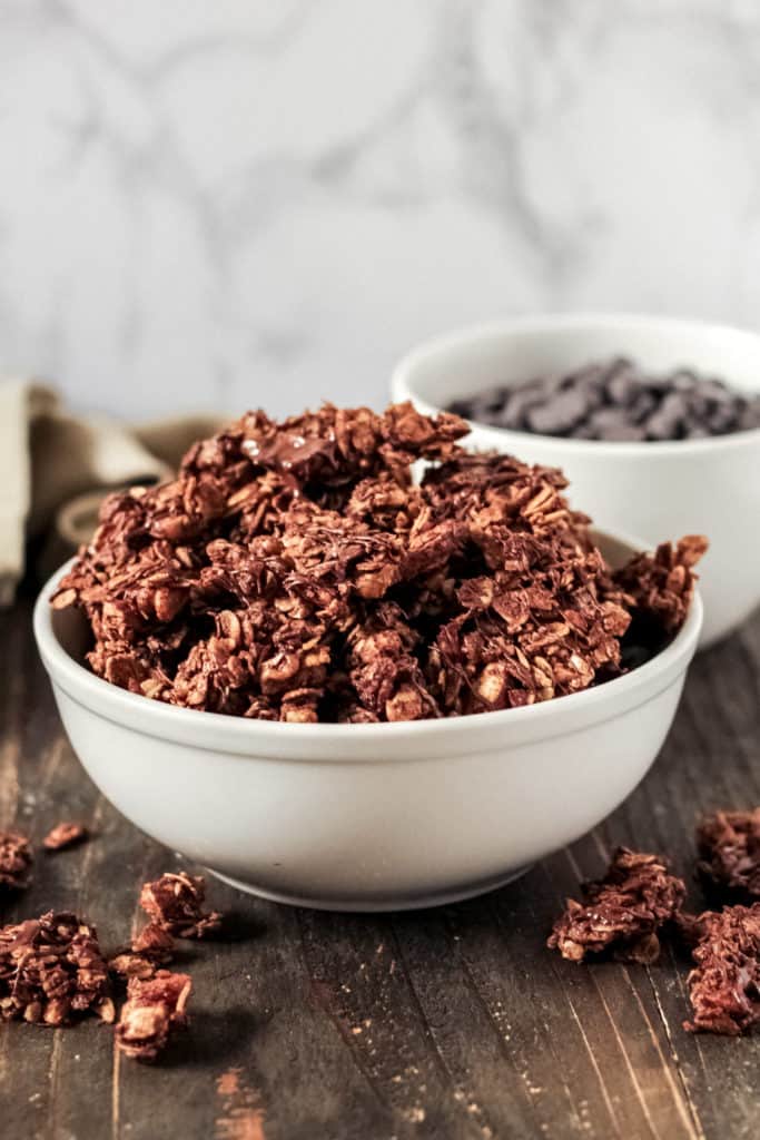 A bowl of chocolate granola with a bowl of chocolate chips behind