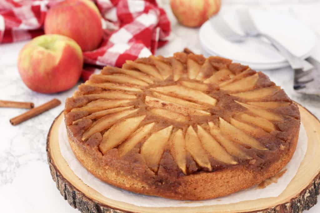 Cake on a wood platter with apples and cinnamon sticks around.